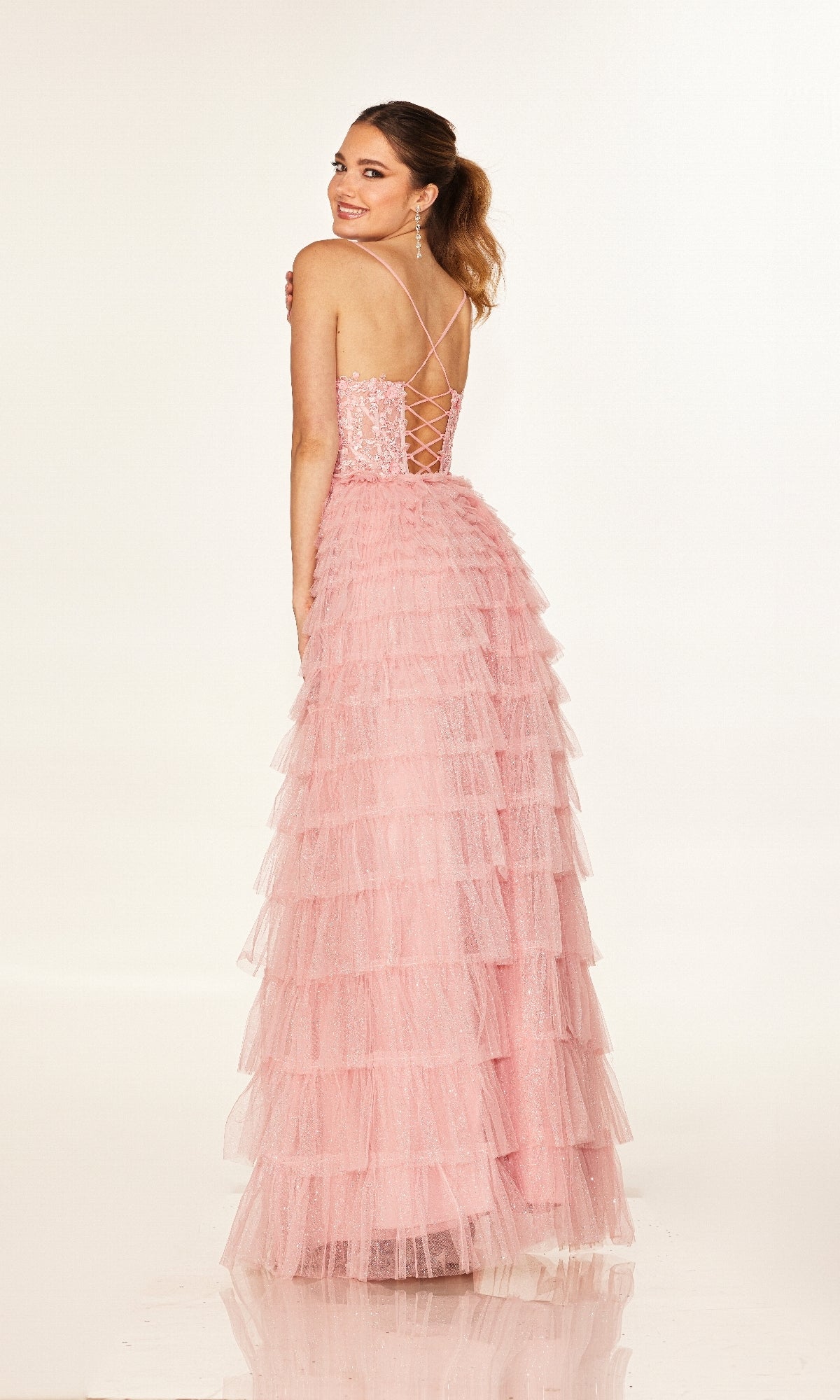 A woman poses, looking over her shoulder in the Cecilia Couture 2613 long prom dress—a pink, tiered tulle gown with a lace-up open back and embroidered corset bodice.